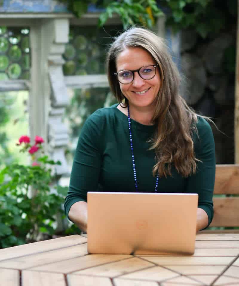 Mareile Poettering sitzt mit Laptop an einem Holztisch in ruhiger Gartenszene. Psychologin für Selbstverbindung und Einzelbegleitung.